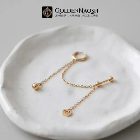 A pair of gold body jewelry pieces, including a ring with a dangling chain and bead, and a barbell with a dangling chain and small charm, displayed on a textured white ceramic dish. The brand logo 'GOLDENNAQSH' is visible at the top.