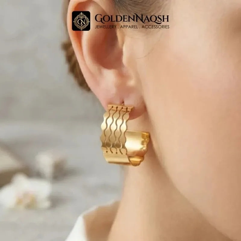A close-up of a person's ear wearing a wide, gold-toned hoop earring with wavy cutout details. The brand logo 'GOLDENNAQSH' is visible at the top of the image.