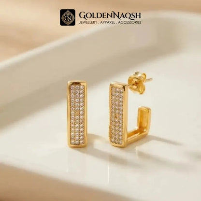 A pair of gold rectangular hoop earrings with rows of small clear pavé stones, displayed on a white tray. The brand logo 'GOLDENNAQSH' is visible at the top of the image.
