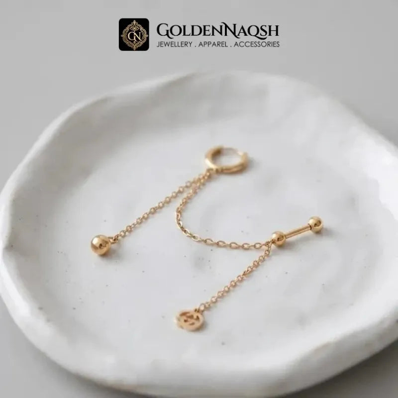A pair of gold body jewelry pieces, including a ring with a dangling chain and bead, and a barbell with a dangling chain and small charm, displayed on a textured white ceramic dish. The brand logo 'GOLDENNAQSH' is visible at the top.