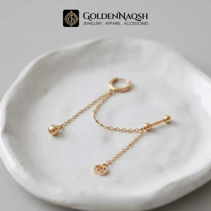 A pair of gold body jewelry pieces, including a ring with a dangling chain and bead, and a barbell with a dangling chain and small charm, displayed on a textured white ceramic dish. The brand logo 'GOLDENNAQSH' is visible at the top.
