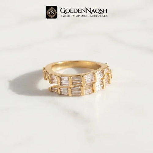 A gold ring with baguette-cut clear stones, displayed on a white marble surface with a soft shadow. The brand logo 'GOLDENNAQSH' is visible at the top of the image.