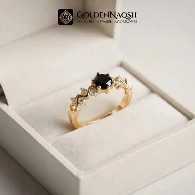 A gold ring with a central black oval stone and small clear accent stones, displayed inside a white jewelry box. The brand logo 'GOLDENNAQSH' is visible at the top of the image.