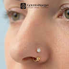 Close-up of a person's nose wearing two small gold nose studs, one with a clear gemstone and one plain, with the GoldenNaqsh brand logo at the top.
