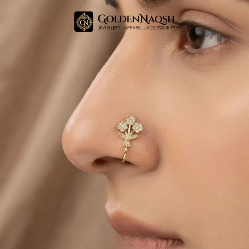 A close-up of a model's nose wearing a delicate gold nose stud with a small floral or leaf-like design set with clear stones. The brand logo 'GOLDENNAQSH' is visible at the top of the image.