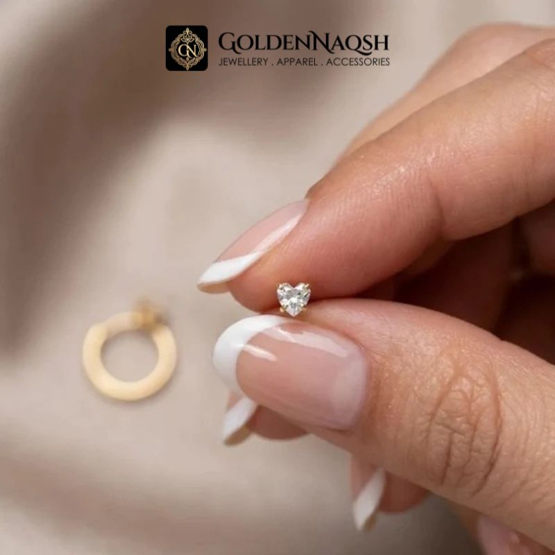 A close-up of a hand with French manicure holding a tiny heart-shaped silver stud earring with a clear gemstone, with a larger gold hoop earring blurred in the background. The GoldenNaqsh brand logo is visible at the top.