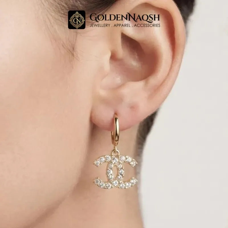 A gold hoop earring with a dangling interlocking CC logo charm encrusted with clear rhinestones, shown worn on a person's ear. The GoldenNaqsh brand logo is visible at the top.