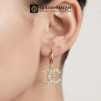 A gold hoop earring with a dangling interlocking CC logo charm encrusted with clear rhinestones, shown worn on a person's ear. The GoldenNaqsh brand logo is visible at the top.