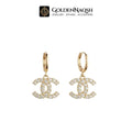 A pair of gold hoop earrings featuring a dangling interlocking CC logo paved with clear crystals, shown against a plain white background with the GoldenNaQSH brand logo at the top.