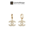 A pair of gold hoop earrings featuring a dangling interlocking CC logo paved with clear crystals, shown against a plain white background with the GoldenNaQSH brand logo at the top.