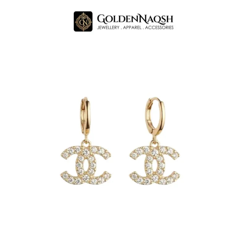 A pair of gold hoop earrings featuring a dangling interlocking CC logo paved with clear crystals, shown against a plain white background with the GoldenNaQSH brand logo at the top.