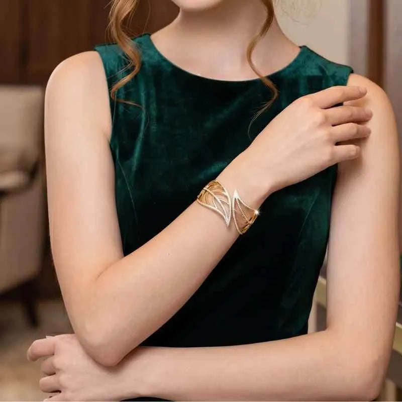 A woman wearing a sleeveless dark green velvet top and a gold geometric cuff bracelet with a silver accent, shown in a softly lit indoor setting.