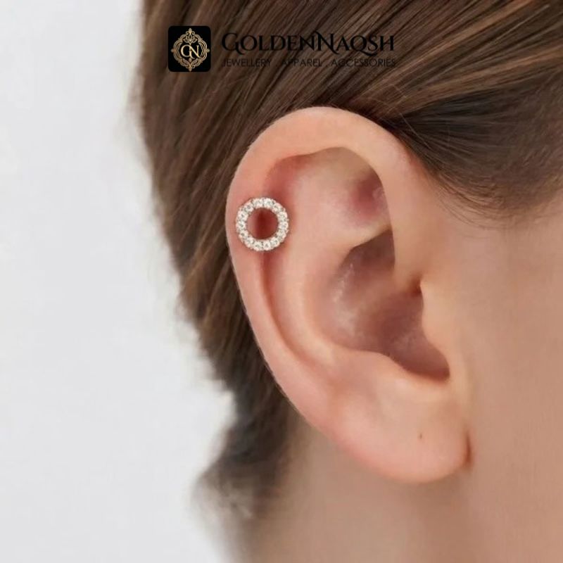 A close-up of a model's ear wearing a small gold hoop earring set with clear stones. The brand logo 'GOLDENNAOOSH' is visible at the top of the image.