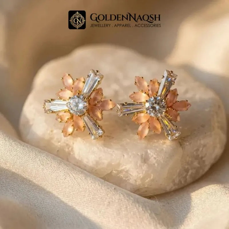 A pair of gold-toned floral stud earrings with peach-pink petals and clear baguette crystals, displayed on a smooth white stone with soft beige fabric. The brand logo 'GOLDENNAQSH' is visible at the top.