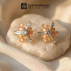 A pair of gold-toned floral stud earrings with peach-pink petals and clear baguette crystals, displayed on a smooth white stone with soft beige fabric. The brand logo 'GOLDENNAQSH' is visible at the top.