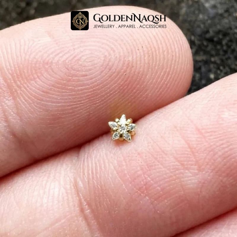 A tiny gold flower-shaped stud earring with clear stones, shown resting on a person's fingers for scale. The GoldenNaqsh brand logo is visible at the top.
