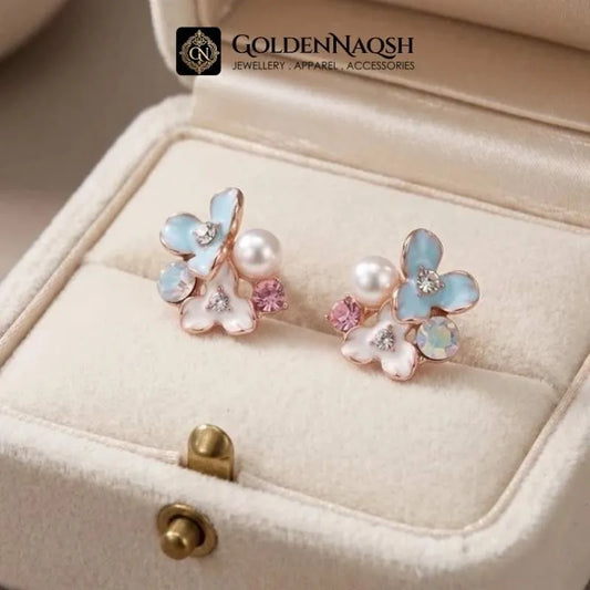 A pair of floral stud earrings displayed in a cream-colored jewelry box. The earrings feature light blue and white enamel flower petals with rose gold metal settings, small white pearls, pink rhinestones, and clear crystal accents.