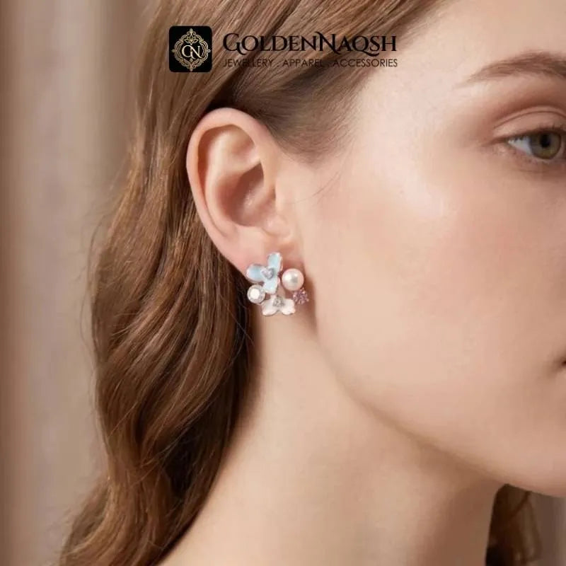 A close-up of a woman's ear wearing a delicate floral stud earring with white pearls and light blue crystals. The brand logo 'GOLDENNAQSH' is visible at the top of the image.