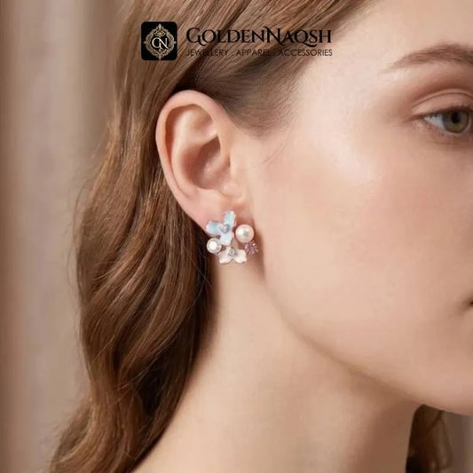 A close-up of a woman's ear wearing a delicate floral stud earring with white pearls and light blue crystals. The brand logo 'GOLDENNAQSH' is visible at the top of the image.