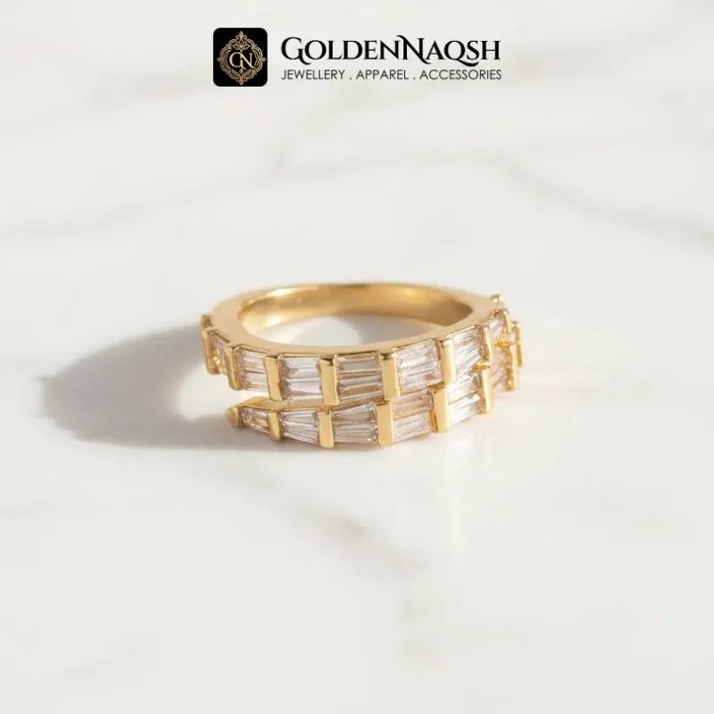 A gold ring with baguette-cut clear stones, displayed on a white marble surface with a soft shadow. The brand logo 'GOLDENNAQSH' is visible at the top of the image.