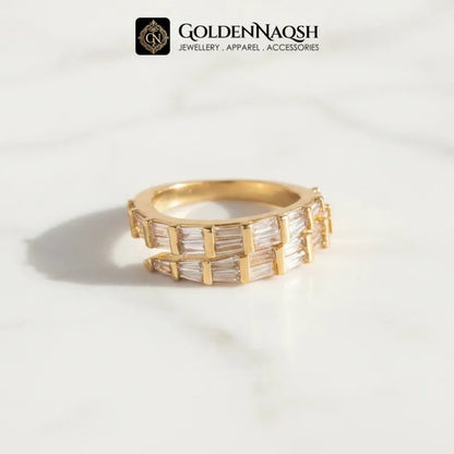 A gold ring with baguette-cut clear stones, displayed on a white marble surface with a soft shadow. The brand logo 'GOLDENNAQSH' is visible at the top of the image.