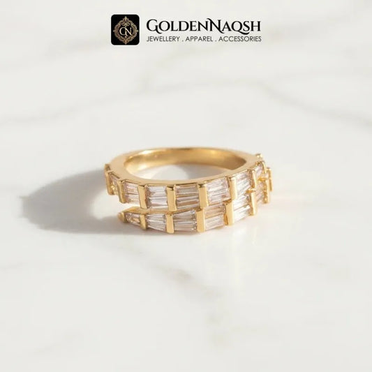 A gold ring with baguette-cut clear stones, displayed on a white marble surface with a soft shadow. The brand logo 'GOLDENNAQSH' is visible at the top of the image.