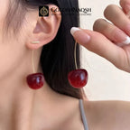 A close-up lifestyle shot of a model wearing a gold post with a glossy deep red cherry drop earring, while holding the matching earring in her hand. The brand logo 'GOLDENNAQSH' is visible at the top.
