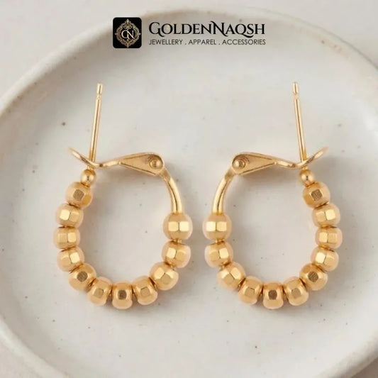 A pair of gold hoop earrings with faceted bead detailing, displayed on a light beige ceramic plate. The brand logo 'GOLDENNAQSH' is visible at the top of the image.